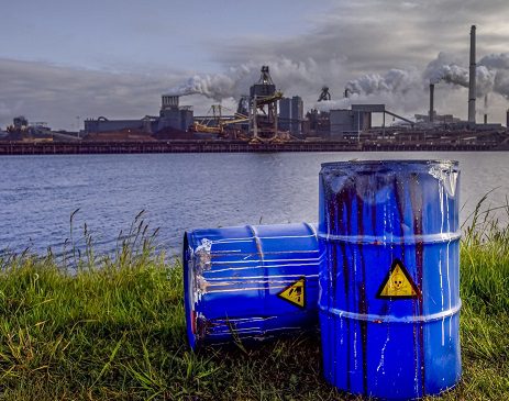 Empty Blue Chemical Waste Drums Lying on an Abandoned Bank with a view on Smoking Exhaust Pipes of a Heavy Industrial Factory